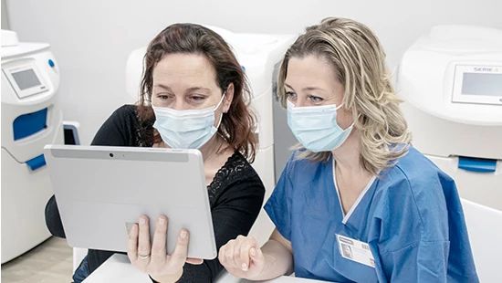 Two people in a medical setting reviewing a tablet, with equipment in the background and faces blurred for privacy.