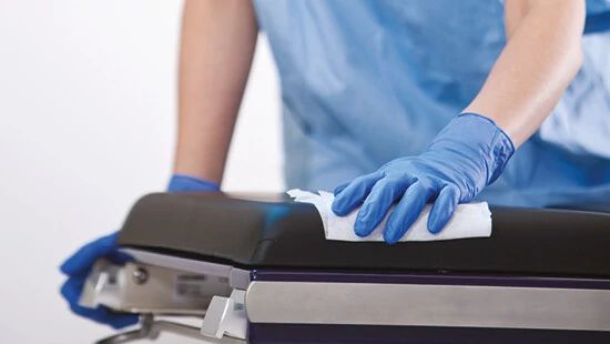 Gloved hand in blue scrubs cleaning a black medical examination table with a white cloth.