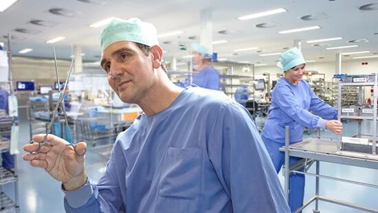 Healthcare professionals in blue scrubs and green caps working in a sterile, well-lit medical room; one holds surgical scissors, others handle instruments.