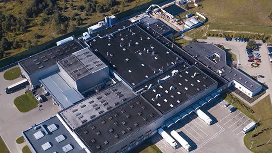 Aerial view of a large industrial facility with interconnected buildings, parking lots, trucks, and surrounding greenery.